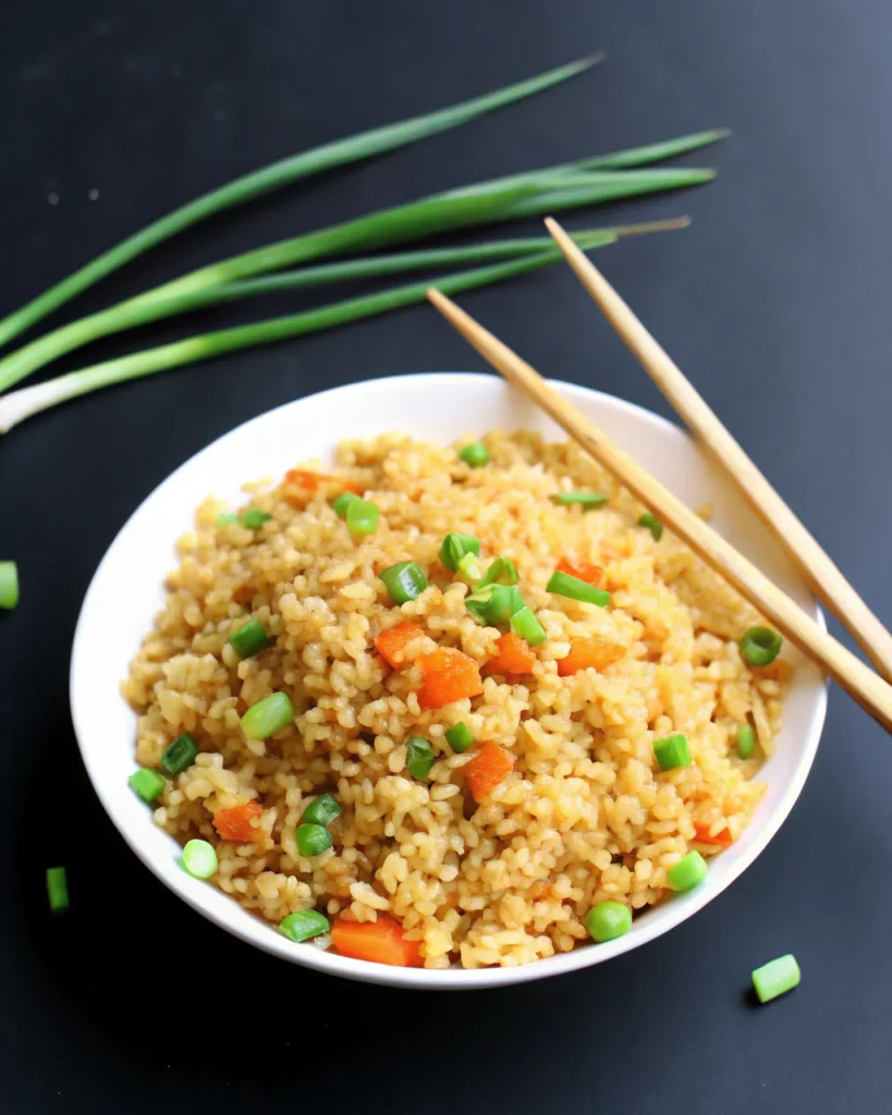 A bowl of Chinese fried rice featuring fluffy rice grains, green peas, diced carrots, and chopped green onions, served with wooden chopsticks. This Chinese fried rice recipe is a classic Asian dish, perfect for a quick and healthy meal.