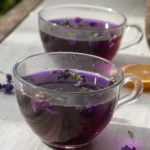 Two clear glass teacups filled with vibrant purple lavender tea, garnished with fresh lavender blossoms and green herbs.