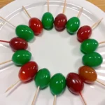 A circular arrangement of glossy red and green candy-coated fruits on toothpicks placed on a white plate.