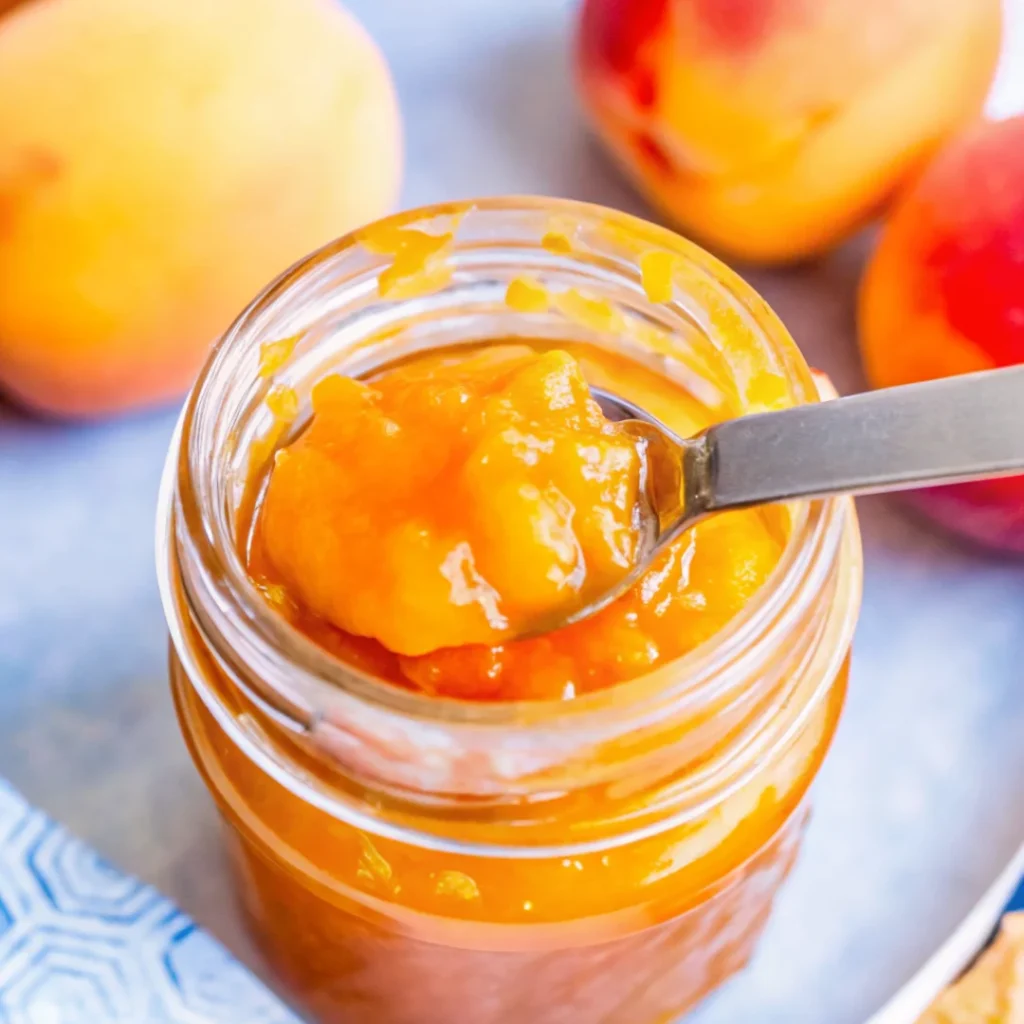 Homemade peach preserves recipe in a glass jar with a spoon, surrounded by fresh peaches.