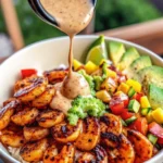 A vibrant shrimp bowl featuring grilled shrimp, sliced avocado, mango salsa, fresh vegetables, and rice, topped with a creamy lime-chili sauce being drizzled over the bowl.