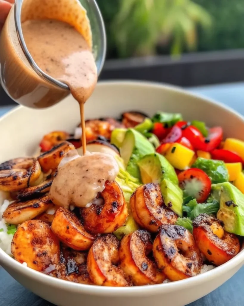 A vibrant shrimp bowl featuring grilled shrimp, sliced avocado, mango salsa, fresh vegetables, and rice, topped with a creamy lime-chili sauce being drizzled over the bowl.