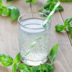 Basil seed drink in clear glass with green striped straw and fresh basil leaves on wooden background