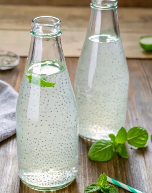 Basil seed drink in clear glass with green striped straw and fresh basil leaves on wooden background
