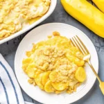 Baked yellow squash casserole with a golden, buttery cracker topping in a white dish, surrounded by fresh bananas and a wooden spoon.