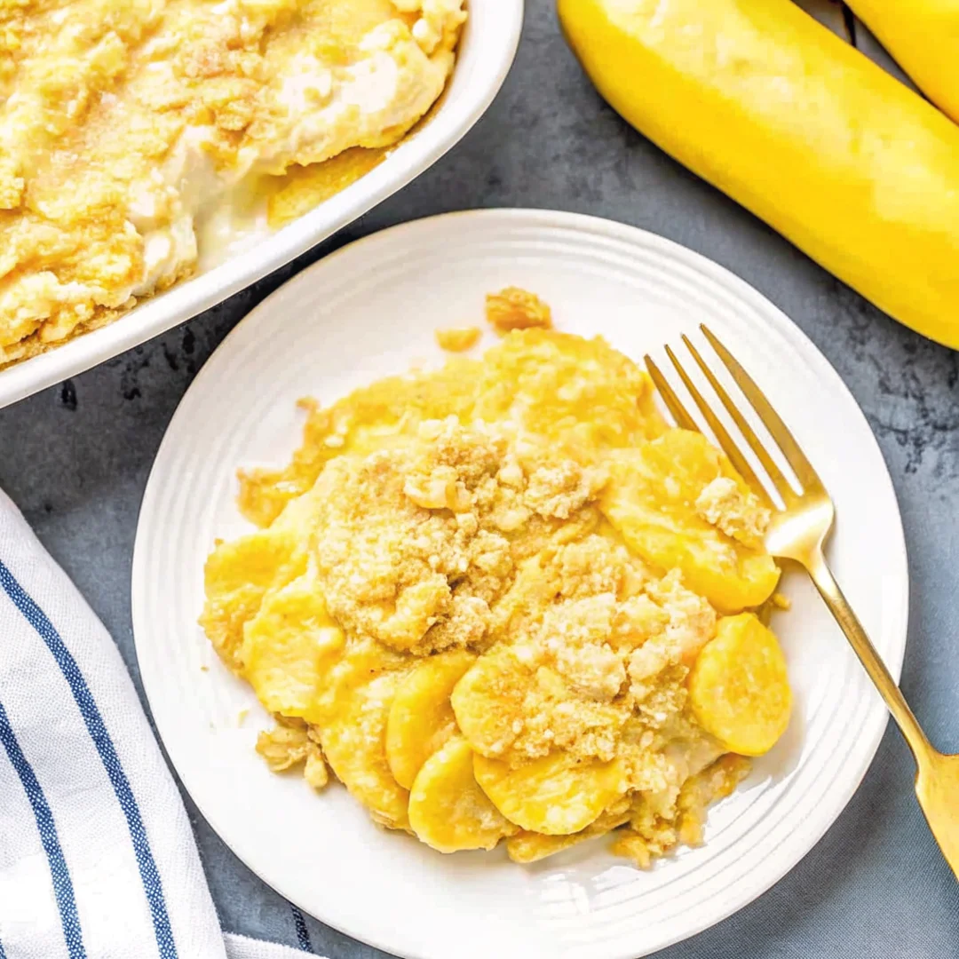 Baked yellow squash casserole with a golden, buttery cracker topping in a white dish, surrounded by fresh bananas and a wooden spoon.