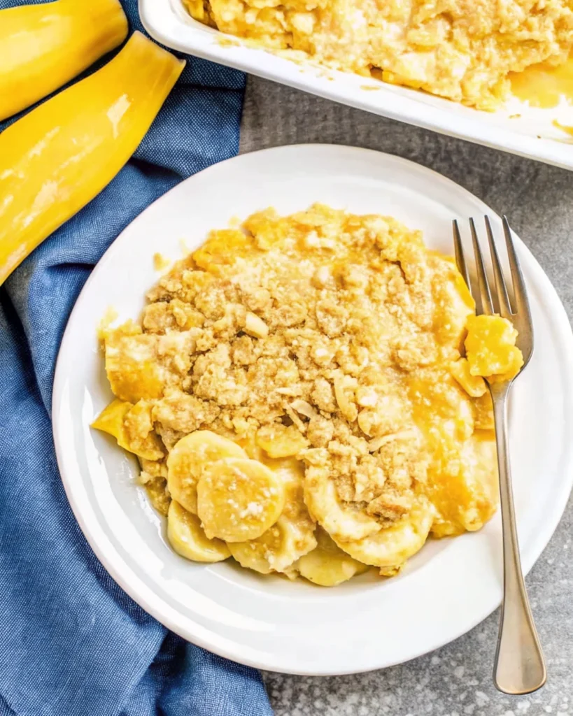 Baked yellow squash casserole with a golden, buttery cracker topping in a white dish, surrounded by fresh bananas and a wooden spoon.