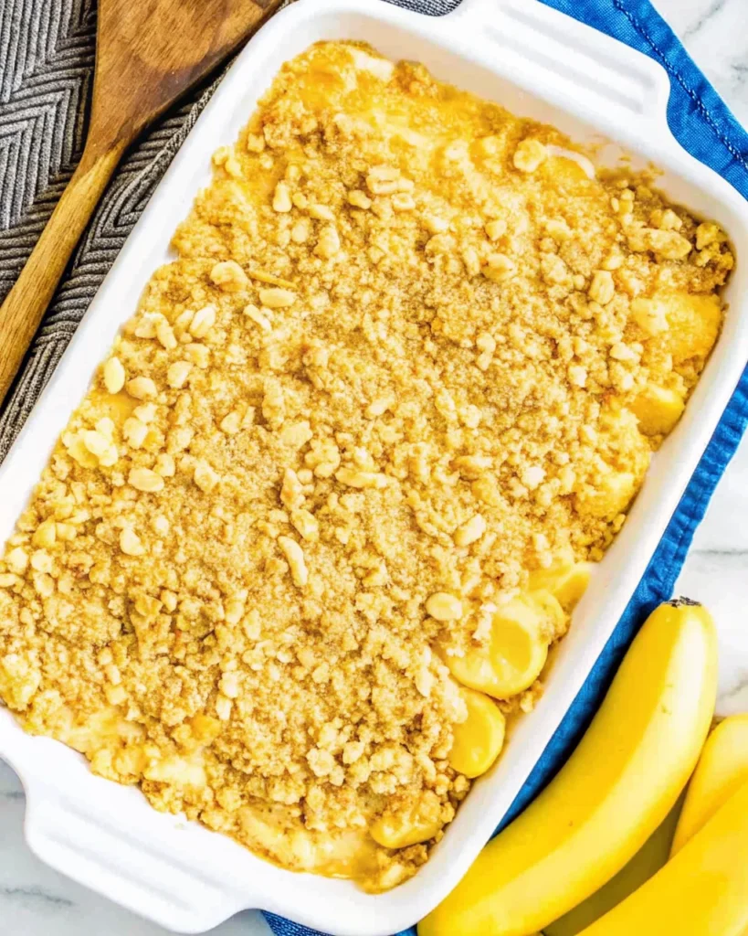Baked yellow squash casserole with a golden, buttery cracker topping in a white dish, surrounded by fresh bananas and a wooden spoon.