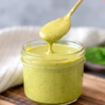 A spoon drizzling creamy balsamic basil vinaigrette into a small mason jar on a wooden surface, with fresh basil leaves in the background.