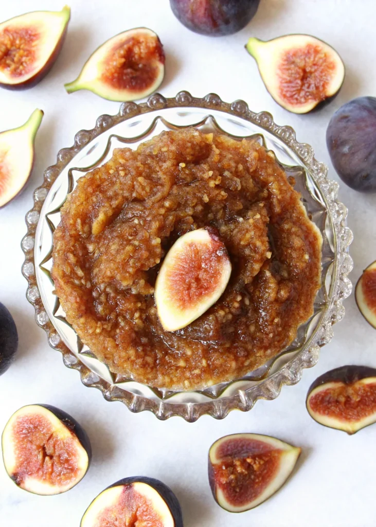 Homemade fig jam made from dried figs in a glass bowl, garnished with fresh fig slices and surrounded by whole figs and yellow flowers.
