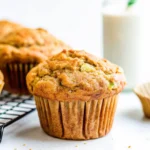 Fluffy whole wheat zucchini carrot muffin with a golden-brown top, set on a marble surface with a cooling rack in the background.