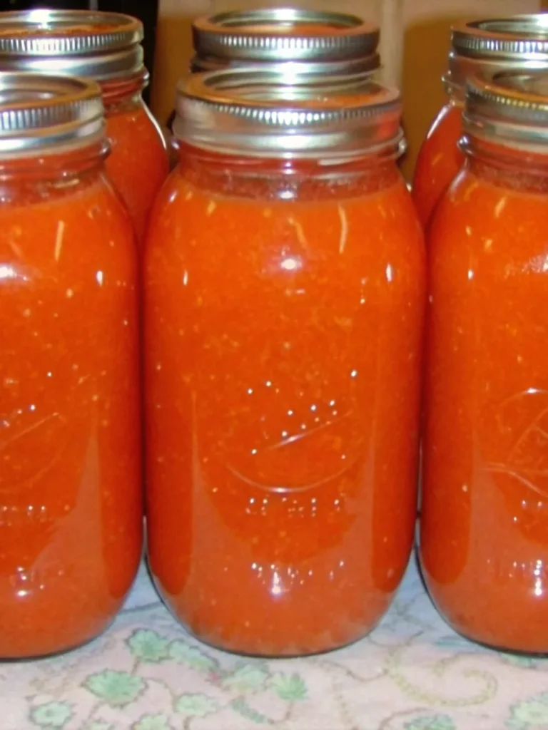 Jars of home-canned crushed tomatoes with metal lids are lined up on a wooden surface in front of a large black canning pot, illustrating the process of canning tomatoes for beginners.