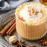 Homemade pumpkin spice latte in clear glass mug with whipped cream being drizzled with honey, surrounded by cinnamon sticks and autumn spices