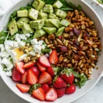 A vibrant strawberry crunch salad for summer eating featuring fresh strawberries, avocado, arugula, goat cheese, candied nuts, seeds, and a tangy champagne vinaigrette, all artfully arranged in a white bowl.