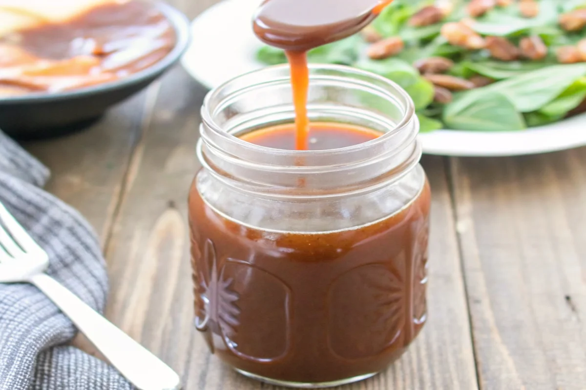 balsamic vinaigrette being drizzled from wooden spoon into glass jar with fresh salad greens in background