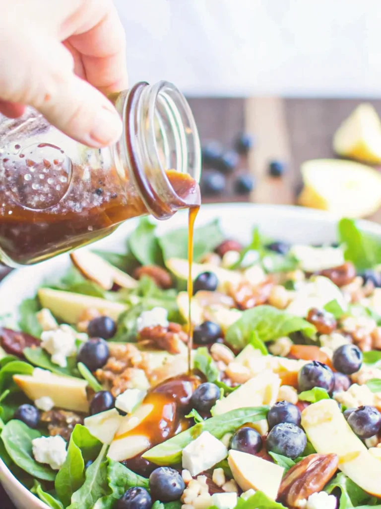 balsamic vinaigrette being drizzled from wooden spoon into glass jar with fresh salad greens in background