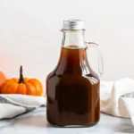 A glass bottle filled with homemade pumpkin spice syrup, surrounded by small pumpkins and kitchen towels, perfect for autumn-inspired recipes.