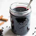 Dark purple elderberry syrup in a glass Mason jar with a spoon and cinnamon sticks scattered nearby