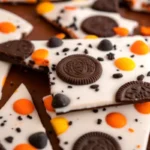Festive Halloween white chocolate bark topped with Halloween Oreos, Reese’s Pieces, and colorful orange and black sprinkles, arranged in broken pieces on a baking sheet.