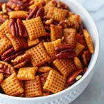 A white bowl filled with Pumpkin Spice Chex Mix, featuring golden Chex cereal squares, pecan halves, and walnut pieces coated in a spiced, buttery mixture for a crunchy autumn snack.