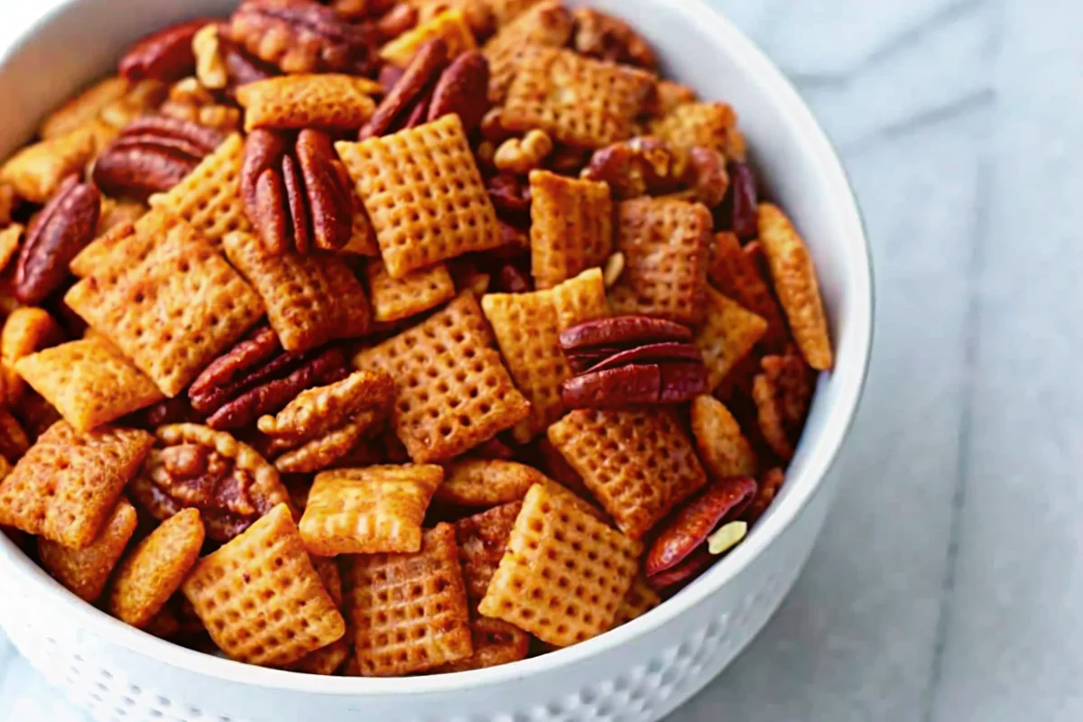 A white bowl filled with Pumpkin Spice Chex Mix, featuring golden Chex cereal squares, pecan halves, and walnut pieces coated in a spiced, buttery mixture for a crunchy autumn snack.