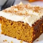 A moist slice of pumpkin spice cake topped with creamy cream cheese frosting and a sprinkle of cinnamon, served on a white plate.