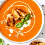 A bowl of roasted tomato soup with fresh tomatoes, topped with crispy grilled cheese croutons, basil, and a swirl of cream.