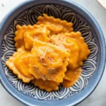 Creamy pumpkin ravioli in rich orange sauce topped with fresh rosemary and grated Parmigiano cheese in white bowl, perfect fall comfort food dinner recipe