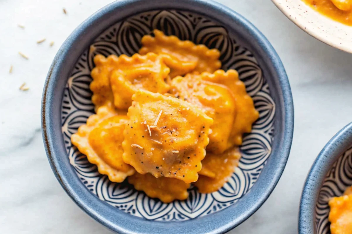 Creamy pumpkin ravioli in rich orange sauce topped with fresh rosemary and grated Parmigiano cheese in white bowl, perfect fall comfort food dinner recipe