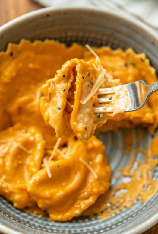 Creamy pumpkin ravioli in rich orange sauce topped with fresh rosemary and grated Parmigiano cheese in white bowl, perfect fall comfort food dinner recipe