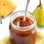 Homemade pear butter recipe in glass jars with fresh Bartlett pears, cinnamon sticks, and warm spices arranged on rustic wooden cutting board, showing the smooth golden-brown texture of the finished pear butter spread