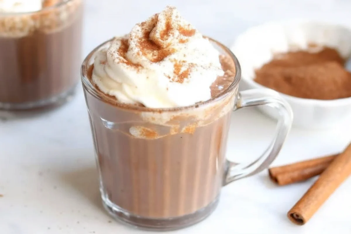 Creamy Pumpkin Spice Hot Chocolate in white mug topped with whipped cream and cinnamon, surrounded by fall decorations and pumpkin spice ingredients