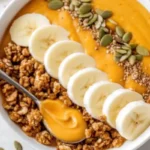 Thick pumpkin smoothie bowl with granola, nuts, and pumpkin seeds arranged artistically in white bowl on marble counter
