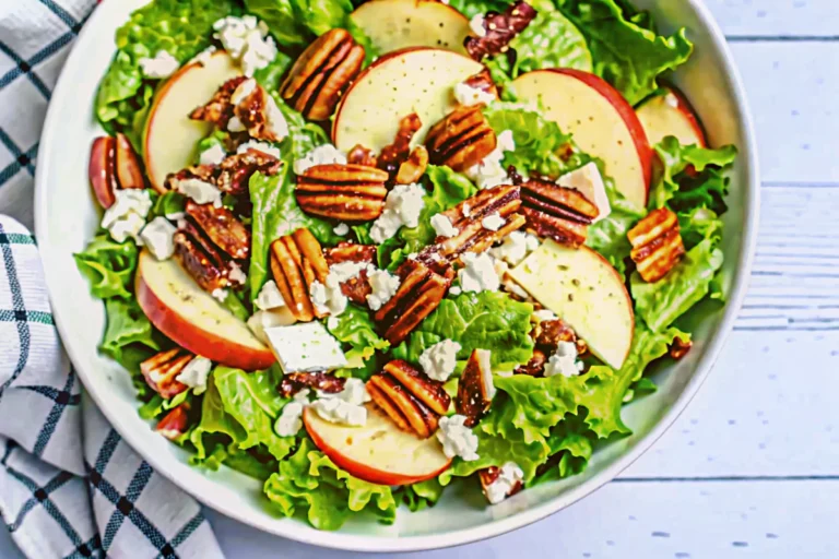 A beautifully composed autumn salad featuring fresh mixed greens as the base, topped with thin slices of crisp Honeycrisp apples, crumbled white feta cheese, golden toasted walnuts, and ruby red dried cranberries. The honey mustard vinaigrette glistens lightly over the ingredients, creating an appetizing display of fall colors and textures. The salad is presented in a clean white serving bowl that highlights the natural colors of this seasonal autumn salad recipe.