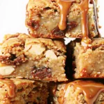A close-up photograph of freshly baked Twix Blondies arranged on a white serving plate, showcasing the golden-brown surface with visible Twix candy pieces, chocolate chips, and a glossy caramel drizzle. The blondies display the perfect fudgy texture with slightly crackled tops and gooey caramel centers, photographed with natural lighting to emphasize the rich, indulgent appearance.