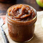 Homemade apple butter in glass jar with fresh apples and cinnamon sticks on rustic wooden background, perfect for fall breakfast spreads